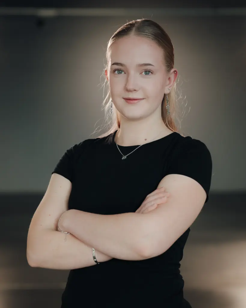 Junge Frau mit zurückgebundenem Haar und schwarzem T-Shirt steht mit verschränkten Armen selbstbewusst vor neutralem Hintergrund.