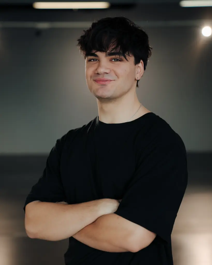 Junger Mann mit dunklen Haaren und schwarzem T-Shirt steht mit verschränkten Armen lächelnd vor neutralem Hintergrund.