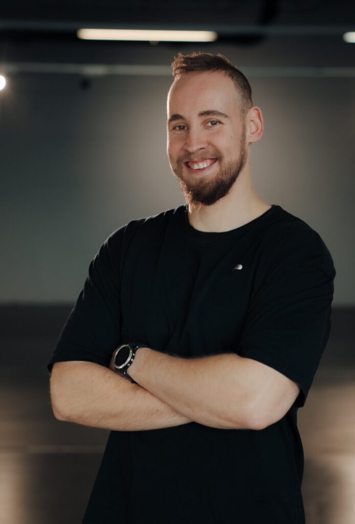 Mann mit dunklem Haar und sportlicher Ausstrahlung steht selbstbewusst mit verschränkten Armen vor neutralem Hintergrund.